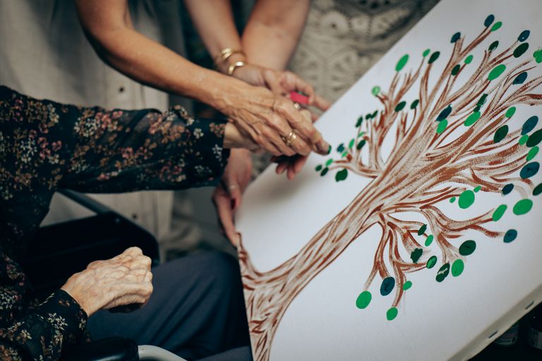 An elderly person and caregiver collaboratively painting a tree art piece, emphasizing creativity and care.